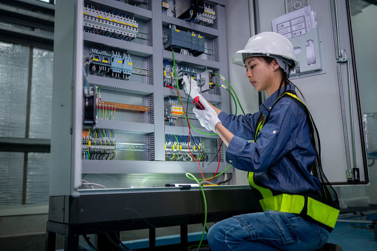 A female technician wearing a hard hat and safety vest works on an open electrical control panel, testing wiring with a multimeter. The image shows industrial electrical infrastructure, technical skills, and hands-on maintenance work in a controlled indoor facility.