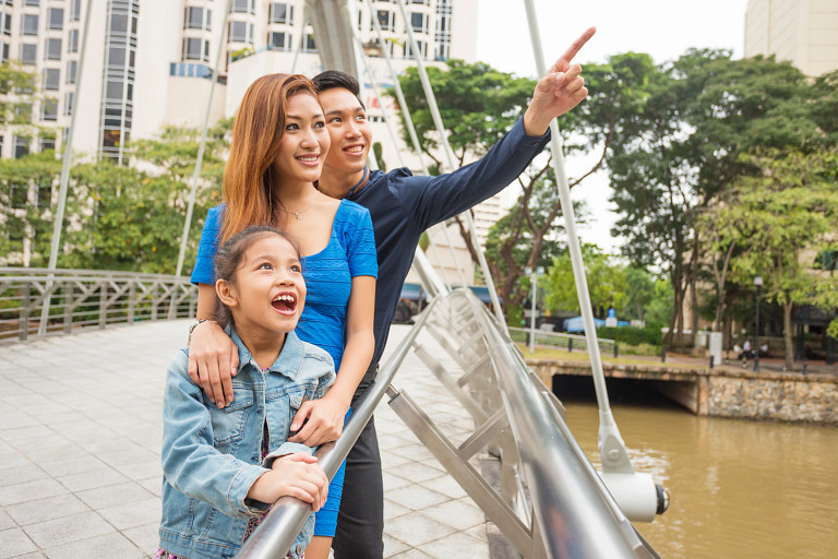 A family of three stands on a pedestrian bridge in a city park, with parents and a young child looking ahead. The father points into the distance while the mother and child smile. The scene depicts an urban lifestyle and family life, with modern buildings, greenery, and a river in the background.
